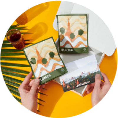 A person holds a bag of tea from Burma in one hand and a postcard in the other, with their Atlas Tea package in the background.