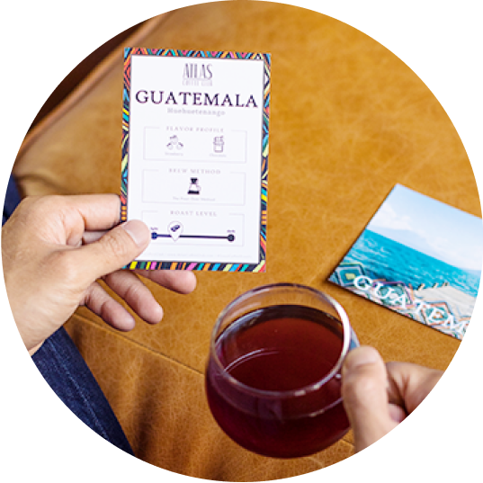 A person holds a cup of coffee in one hand and a card with tasting notes in the other, with a postcard in the background.