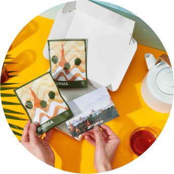 A person holds a postcard and a bag of tea from Burma, with a teapot and cup of red tea in the background on a bright yellow surface.