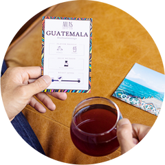 A person holds a cup of coffee in one hand and a card with tasting notes in the other, with a postcard in the background.