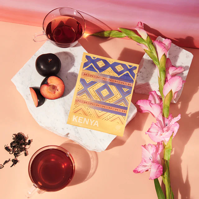A vibrant bag of tea from Kenya is surrounded by plums, pink flowers,  a cup of tea and loose tea leaves on a light pink background.