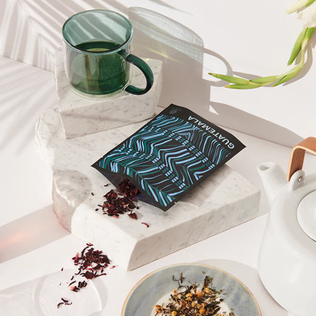 A bag of tea from Guatemala is surrounded by a cup of tea, loose tea leaves and a white tea pot on a white marble surface.