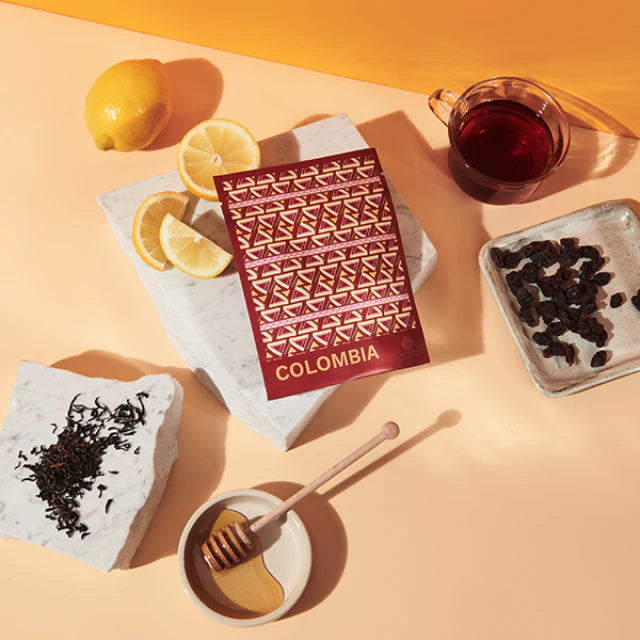 A red bag of tea from Colombia is surrounded by lemon wedges, a cup of tea, loose tea leaves, and a honey comb with honey, all on a light peach background.