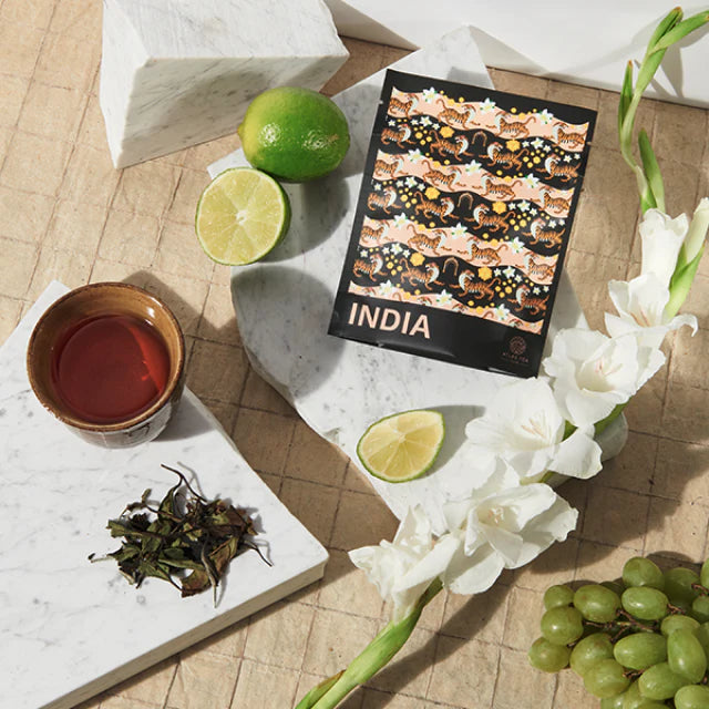 A bag of tea from India sits on a white marble slab surrounded by lime wedges, white flowers, green grapes, a cup of tea and loose tea leaves.