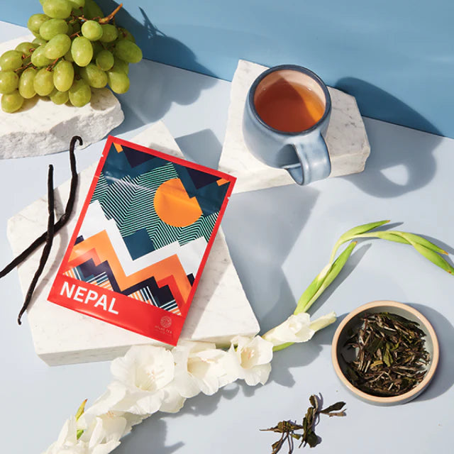 A vibrant bag of tea from Nepal is surrounded by green grapes, a cup of tea, a cup of loose tea leaves and white flowers on a light blue background.